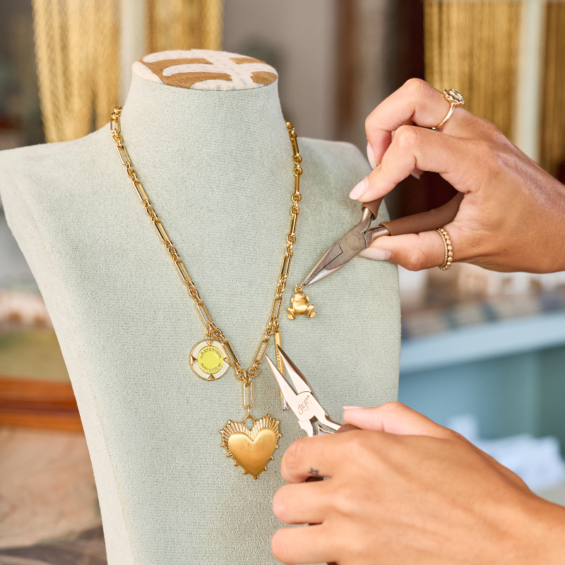 A close-up of a gold necklace with a heart-shaped pendant displayed on a mint green jewelry bust, with a person's hands using pliers to adjust or repair the pendant. The background is a softly blurred indoor setting with warm tones.