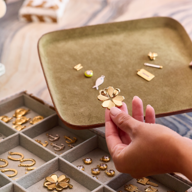 A hand holds a gold four-leaf clover pendant over an open jewelry organizer tray, with a larger olive-green jewelry tray in the background holding other small jewelry pieces.