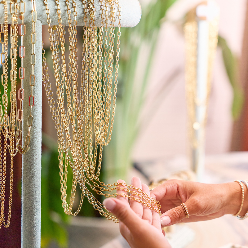 Close-up of a person's hands holding a delicate gold chain necklace, with multiple gold chains hanging from a display stand in a softly blurred boutique setting.
