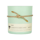 A green soy wax candle with a gold lurex bracelet tied around it, featuring three brass charms, labeled 'HART CHARLESTON CHARM CANDLE'.