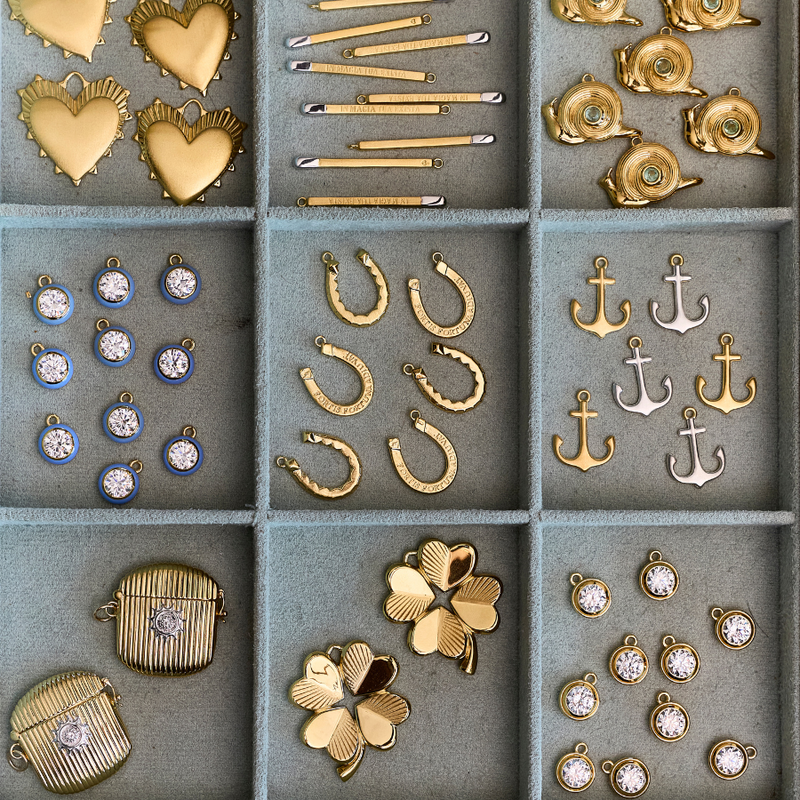 A 3x3 grid of jewelry charms and pendants displayed in a gray divided tray, including hearts, bars, shells, horseshoes, anchors, clovers, and small round charms with clear stones.