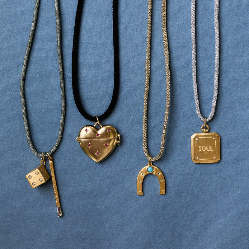 HART Cord Necklaces. Four distinct gold-toned necklaces with unique charms are displayed against a textured blue background. The necklaces include a silver cord with a dice and key pendant, a black cord with a heart-shaped locket, a gold chain with a gold horseshoe pendant featuring a turquoise accent, and a silver chain with a gold square pendant engraved with the word 'SOUL'.
