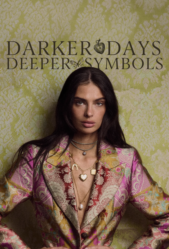 A model wearing a colorful paisley blazer and layered gold necklaces, posed against a green damask wallpaper background with the text 'DARKER DAYS DEEPER SYMBOLS' at the top.