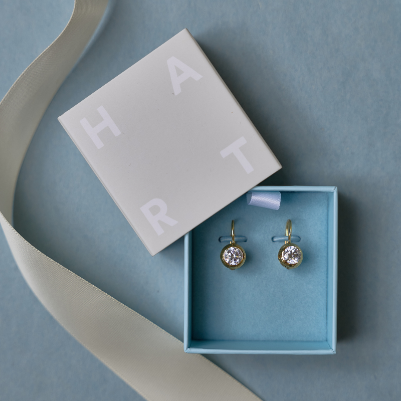 Best gift for brides: A pair of gold hoop earrings with round clear crystal drops, presented in a light blue jewelry box next to a white HART box lid with a white ribbon on a blue background.