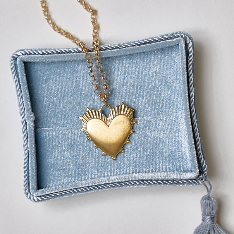 A gold heart pendant necklace with a textured, flame-like top design, resting inside an open light blue velvet jewelry box with a tassel.