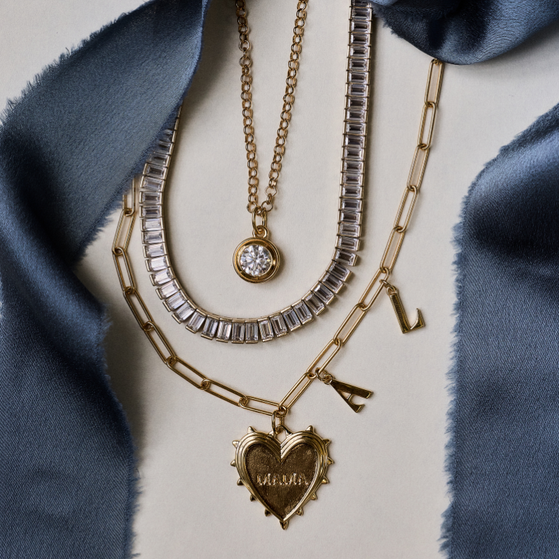 Layered gold necklaces displayed on a light beige surface with a blue satin fabric backdrop. The necklaces include a gold heart pendant engraved with 'MIAHA', a silver-toned rectangular-link chain, a gold chain with a round clear gemstone pendant, and a gold chain with letter charms.