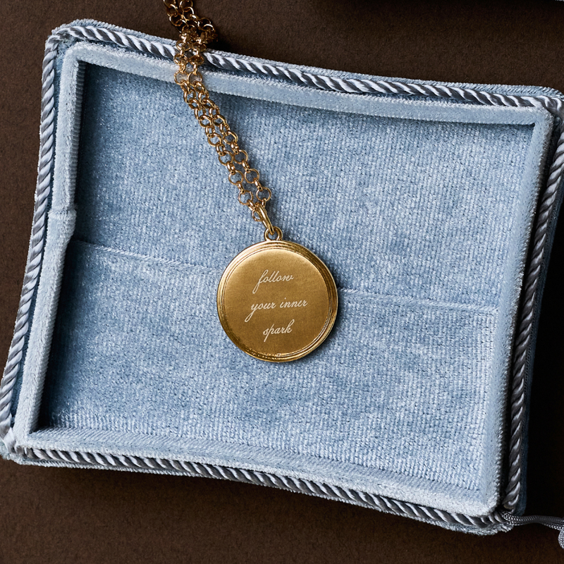 A gold pendant necklace with the engraved words 'follow your inner spark' resting inside an open light blue velvet jewelry box on a dark brown background.