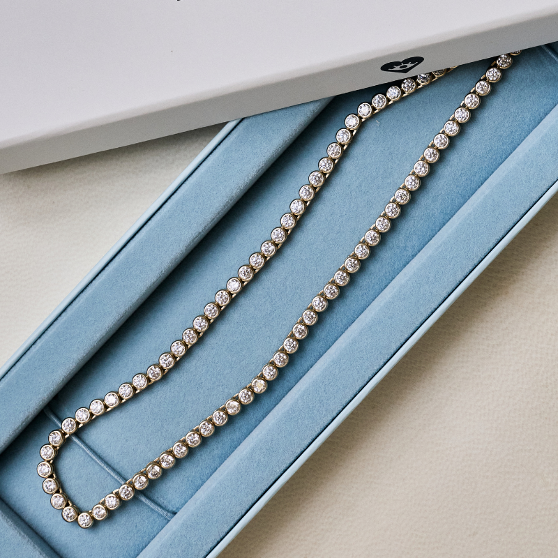 A tennis-style necklace with clear round stones set in a yellow-gold-tone metal, displayed inside an open light blue jewelry box on a beige surface.