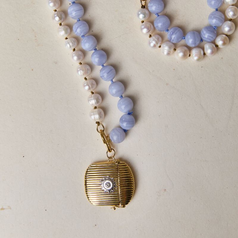 A beaded necklace with white pearl-like beads transitioning into light blue stone beads, featuring a gold-toned round pendant with a small central clear stone, laid on a light beige surface.