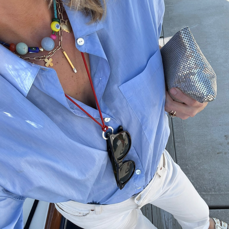 A close-up lifestyle shot of a person wearing a light blue button-down shirt, white pants, and a red cord necklace holding sunglasses. They are also wearing a colorful beaded necklace and holding a silver sequined clutch. wearing HART jewelry 