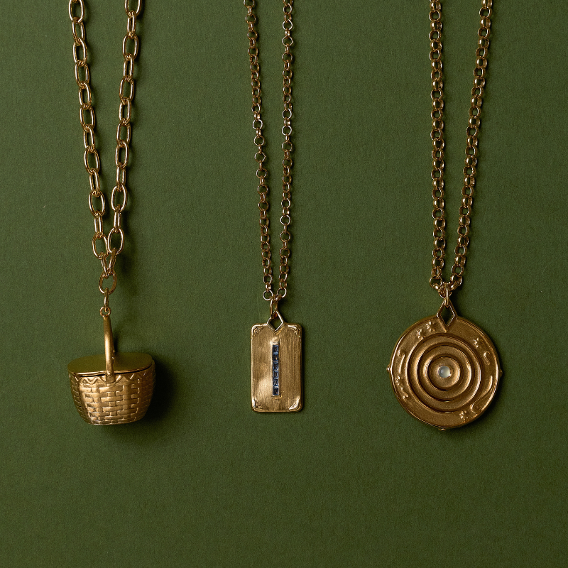 Three gold-toned chain necklaces with distinct pendants are displayed against a solid dark green background. The left pendant is a miniature woven basket, the middle is a rectangular tag with a vertical blue stone accent, and the right is a circular target design with a central white stone.