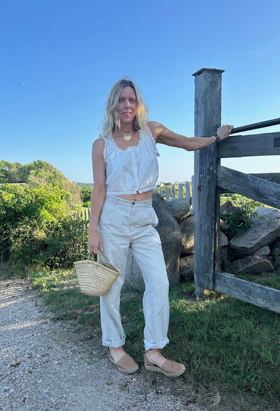 A woman stands outdoors next to a rustic wooden fence, wearing a white sleeveless ruffled crop top and matching white trousers, holding a woven straw bag. wearing HART jewelry 