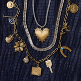 A styled flat-lay of layered gold custom charm necklace stack including smiley face charm, see no evil charm, oyster charm, four leaf clover charm, bezel charm, etched vesta case charm, key charm, horseshoe charm, bird charm, etched heart charm. Layered with a baguette tennis necklace, silver cord with heart of gold charm. 