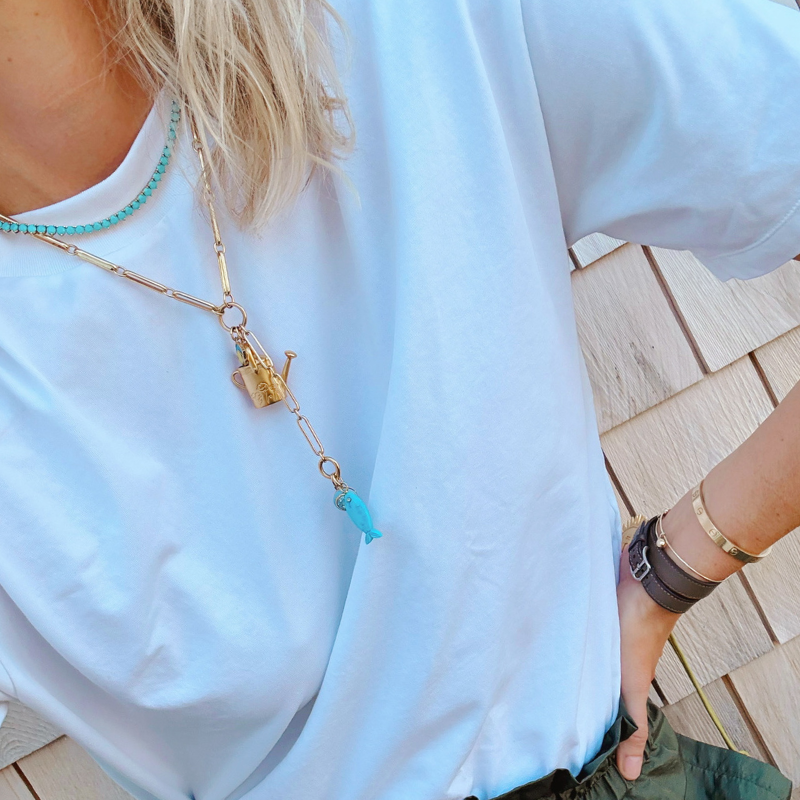 A close-up lifestyle shot of a person wearing a gold-tone chain necklace with a watering can charm and a turquoise fish charm, layered over a white t-shirt.