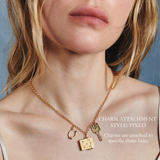 Close-up of a woman wearing a delicate gold chain necklace with three charms: a horseshoe, a small key, and etched vesta case. White text on the right reads 'CHARM ATTACHMENT STYLE: FIXED' and 'Charms are attached to specific chain links.