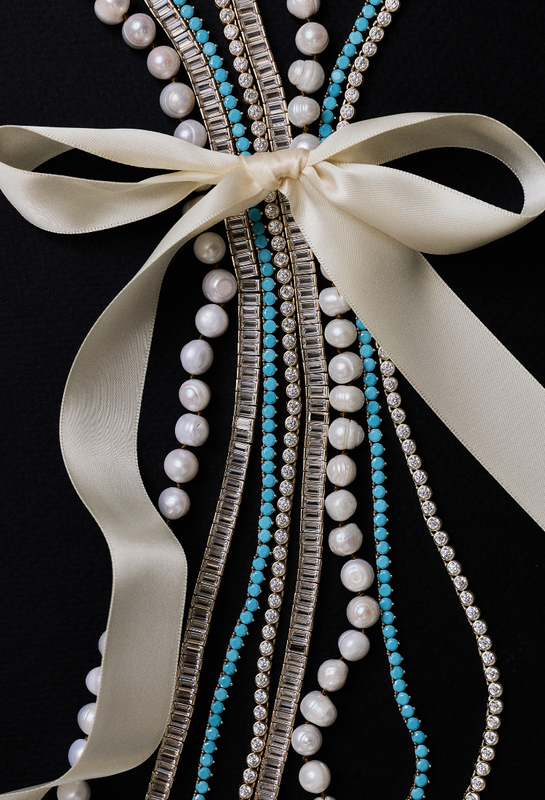 Gift HART jewelry: A close-up of a decorative embellished garment or accessory featuring multiple strands of white pearls, turquoise beads, clear rhinestone-like beads, and a wide cream satin ribbon tied in a bow against a black background.
