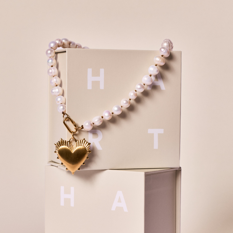 Gift pearl necklace: A pearl necklace with a gold heart-shaped pendant, draped over a beige box with white letters, on a matching beige background.