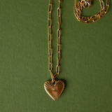 A gold paperclip chain necklace with a textured heart pendant, displayed on a dark green background with a second coiled section of the chain visible in the upper right.