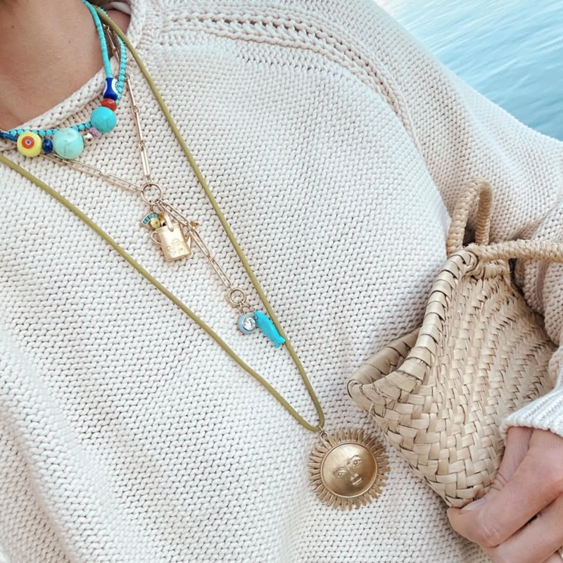 A close-up lifestyle shot of a person wearing a cream knit sweater, layered gold necklaces (one with a sun pendant, one with a small gold  watering can charm, and one with turquoise beads), holding a woven straw bag. wearing HART jewelry 