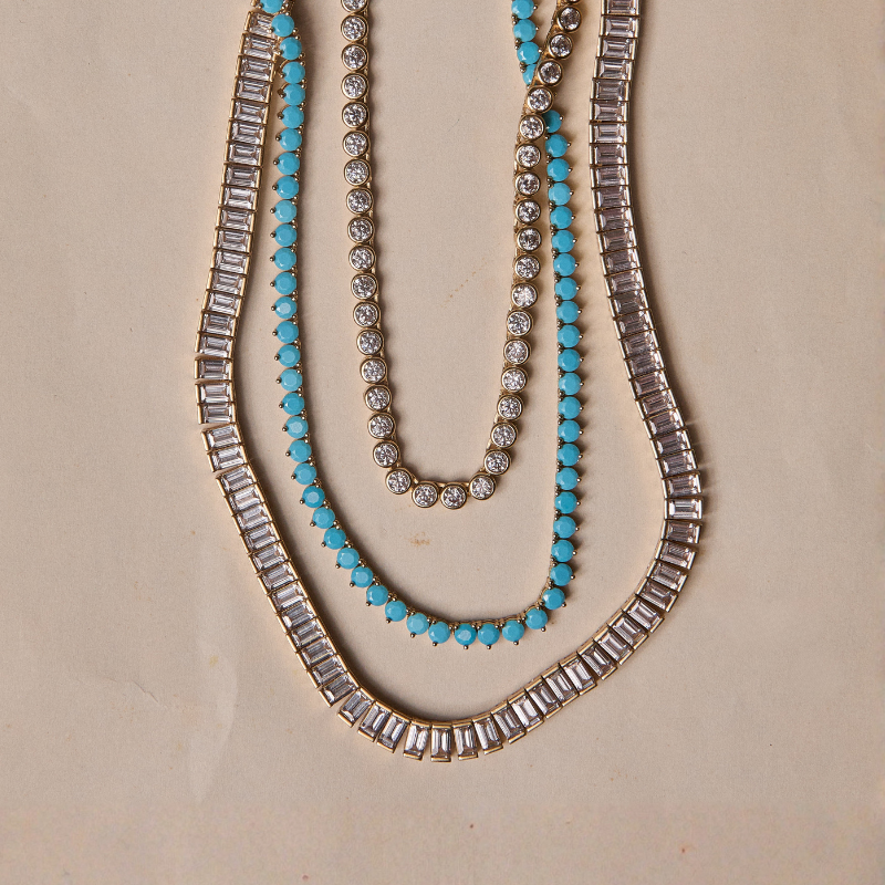 HART Tennis Necklaces: Three gold-toned necklaces arranged concentrically on a beige background: one with clear baguette-cut stones, one with turquoise and clear stones, and one with clear round stones.