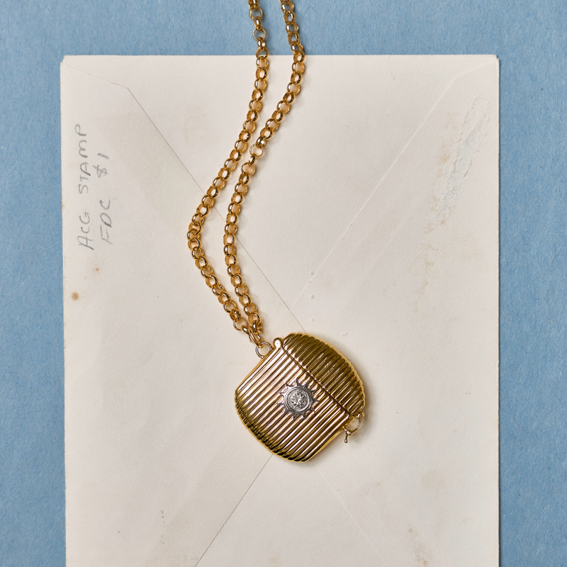HART anniversary gift. The vintage inspired Vesta Case Necklace with a striped pattern and a central stone emblem, displayed on an off-white envelope against a blue background.