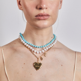 Person wearing a turquoise and pearl beaded necklace with gold heart pendant on a plain background