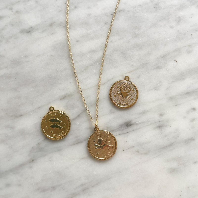 Leo, Circle, zodiac coin is on a dainty gold filled chain. There is a Pisces coin charm and a cancer coin charm in the photo too. All are displayed on a marble countertop.